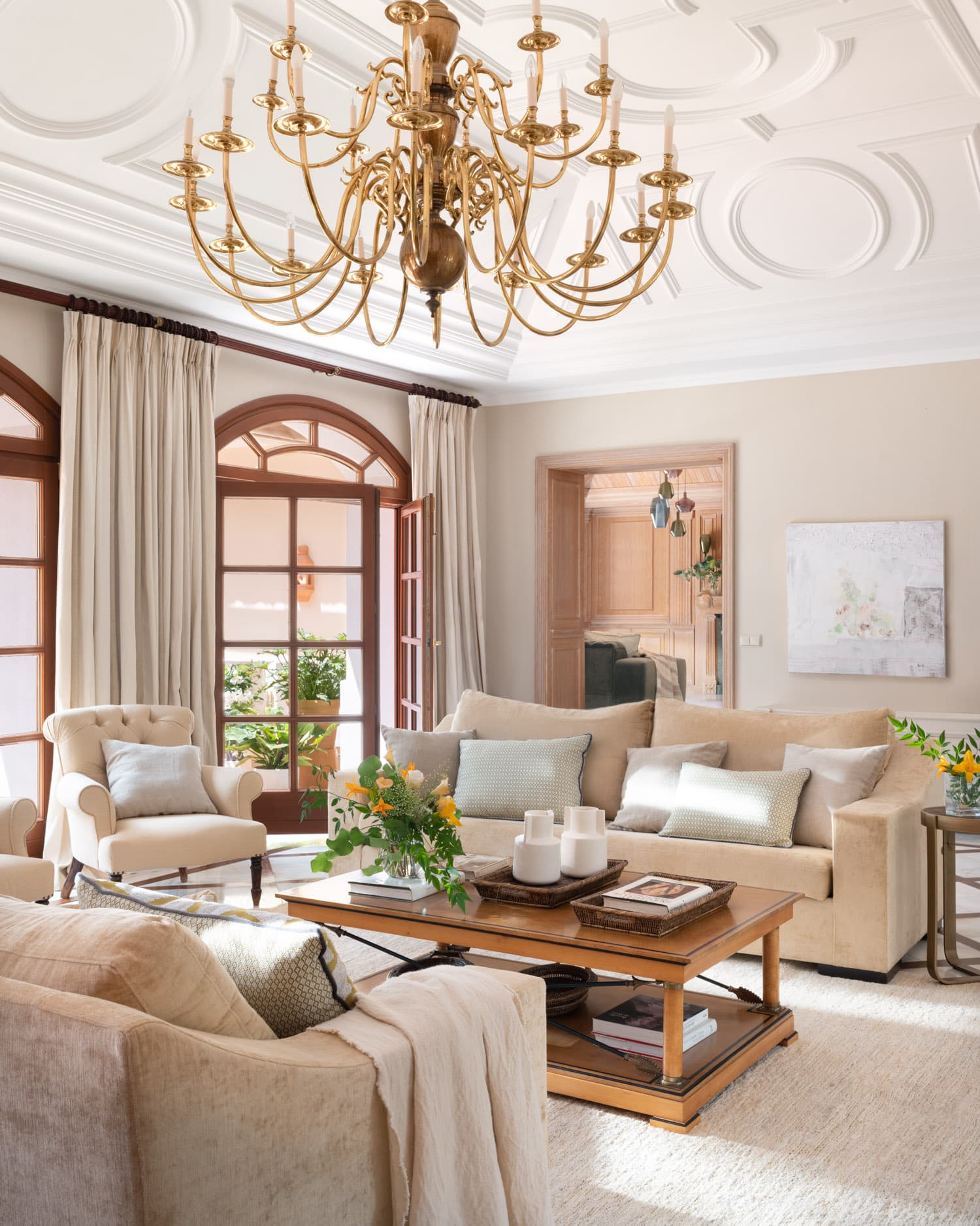 Vista del salón principal con un techo ornamentado de molduras geométricas en blanco y una imponente araña de velas dorada. Los sofás y butacas en tonos crema y beige se agrupan alrededor de una mesa de centro de madera, con la puerta arqueada al fondo revelando una enfilada de estancias. Una obra de arte abstracta en tonos suaves decora la pared lateral.